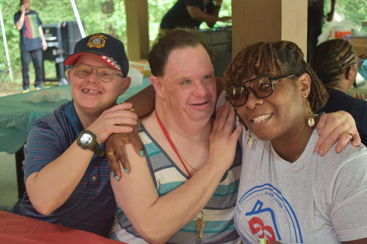 Three adults - two men and a woman sit under a picnic shelter outdoors. They are all smiling and sitting together with their arms around each other .