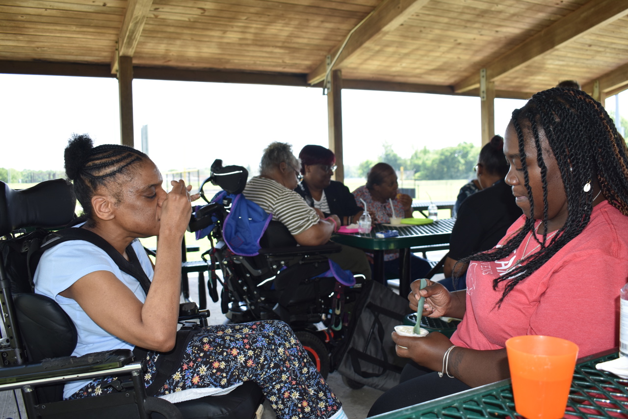 A group of people are sitting together under a covered pavilion, enjoying a meal. In the foreground, a woman in a wheelchair is eating, while another woman across the table stirs her food. In the background, several others are seated at the table, engaging in conversation. The atmosphere appears relaxed, with the group gathered in a peaceful outdoor picnic gathering.