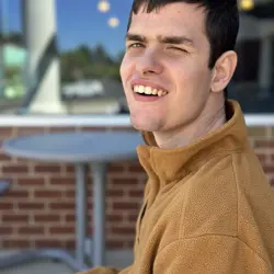 The image shows a young man smiling brightly while sitting outdoors. He is wearing a warm, tan fleece jacket. The background features a brick wall, large glass windows, and outdoor seating, suggesting a café or restaurant setting. The sunlight highlights his face, creating a cheerful and warm atmosphere.