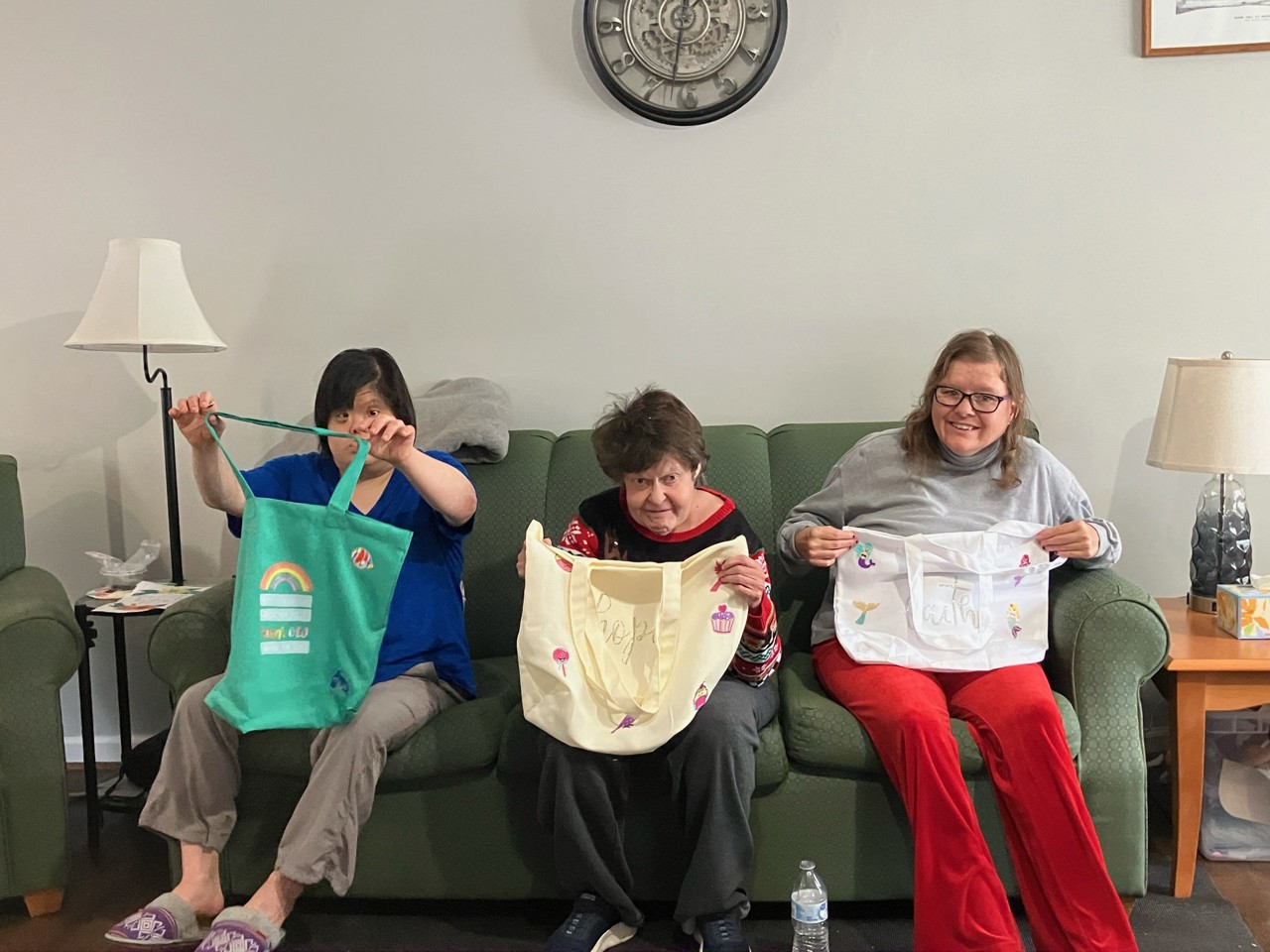 Three women sit on a green couch indoors with a white wall behind them and lamps on either side. They are smiling and showing off their handmade decorated canvas bags. 