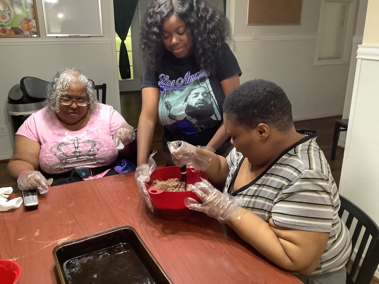 Three women are gathered around a table, preparing food together. The two seated women are wearing gloves and are working with ingredients in a red bowl, while the woman standing is assisting them. The environment suggests a homey and cooperative setting, with everyone focused on the task at hand, creating a sense of teamwork and community.