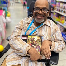 A black man in a brown striped shirt and glasses sits in his wheelchair in the aisle of a store holding a Marvel puzzle box and grinning.