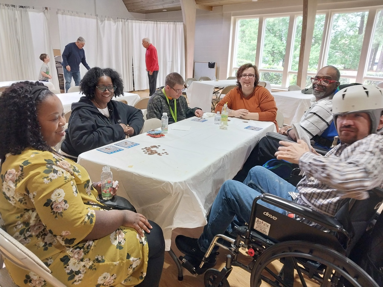 A group of six people sit around a white-covered table in a well-lit room, enjoying a casual gathering. The group includes individuals of diverse ages, with one person wearing a protective helmet and another in a wheelchair. Bingo cards and coins are spread out on the table, suggesting they are playing a game. The atmosphere is relaxed and cheerful, with others walking in the background near large windows that let in natural light. Everyone seems engaged, enjoying each other's company.