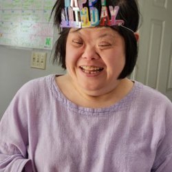 A woman wearing a multicolor Happy Birthday crown and purple shirt is shown closeup with a big smile. Behind her a white interior door and a white board calendar are visible along with a gray wall. 