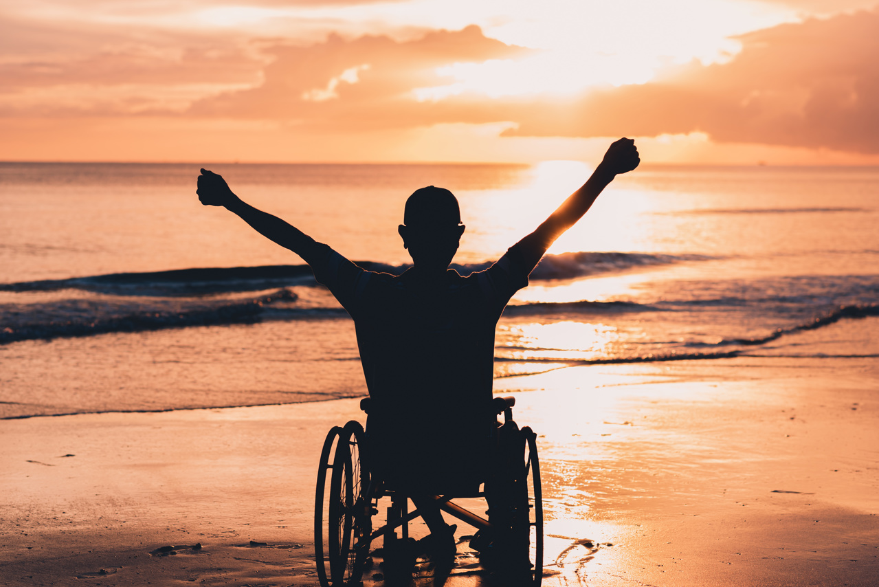 Person in a wheelchair from the back with a silhouette of their shape on the beach looking out toward the waves and the sunset, orange hues