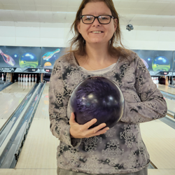 A white woman with shoulder length brown hair and glasses stands smiling in a bowling alley holding a dark purple bowling ball.