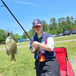 A person is standing outdoors on a sunny day, holding a fishing rod with a small fish on the line. They are wearing a blue and white shirt, dark shorts, a hat, and glasses. Behind him, there is a red folding chair and a grassy area with trees and parked cars in the background.