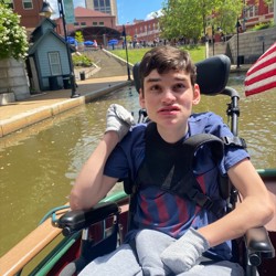 A young man is sitting in a wheelchair on a boat during a canal cruise. He is wearing a navy blue t-shirt, grey pants, and gloves. The background shows a waterway with buildings, trees, and a clear blue sky with a few clouds. An American flag is partially visible on the right side.