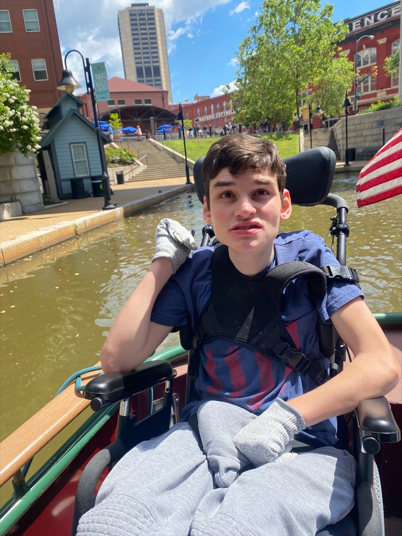 A young man is sitting in a wheelchair on a boat during a canal cruise. He is wearing a navy blue t-shirt, grey pants, and gloves. The background shows a waterway with buildings, trees, and a clear blue sky with a few clouds. An American flag is partially visible on the right side.