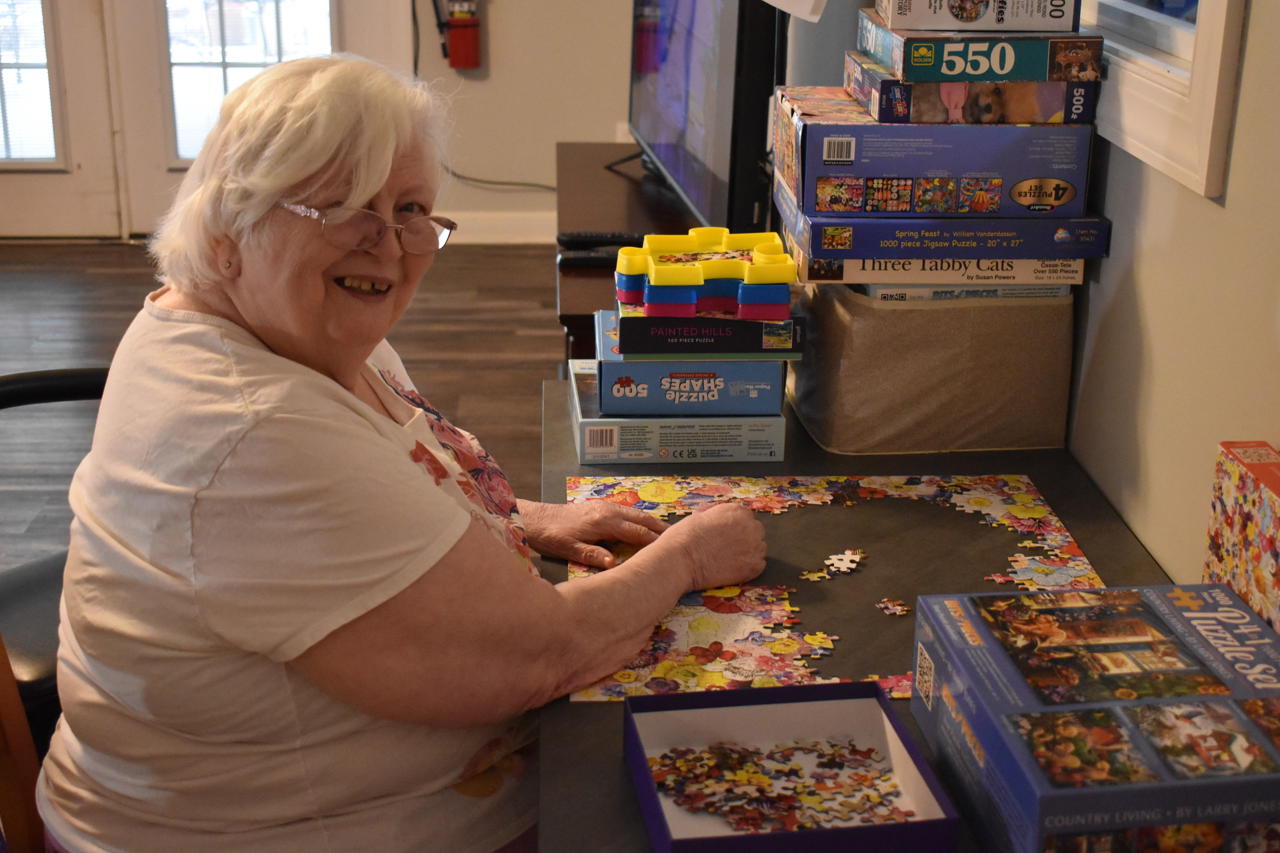 A woman with white hair and glasses sits at a table indoors doing a puzzle, which is complete on the outside edges and still in process in the middle. Other puzzles and games are stacked on the table also. 