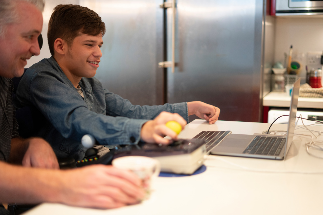 An older man sits behind and to the side of a younger man using an assistive device to access a laptop on the white table in front of them.