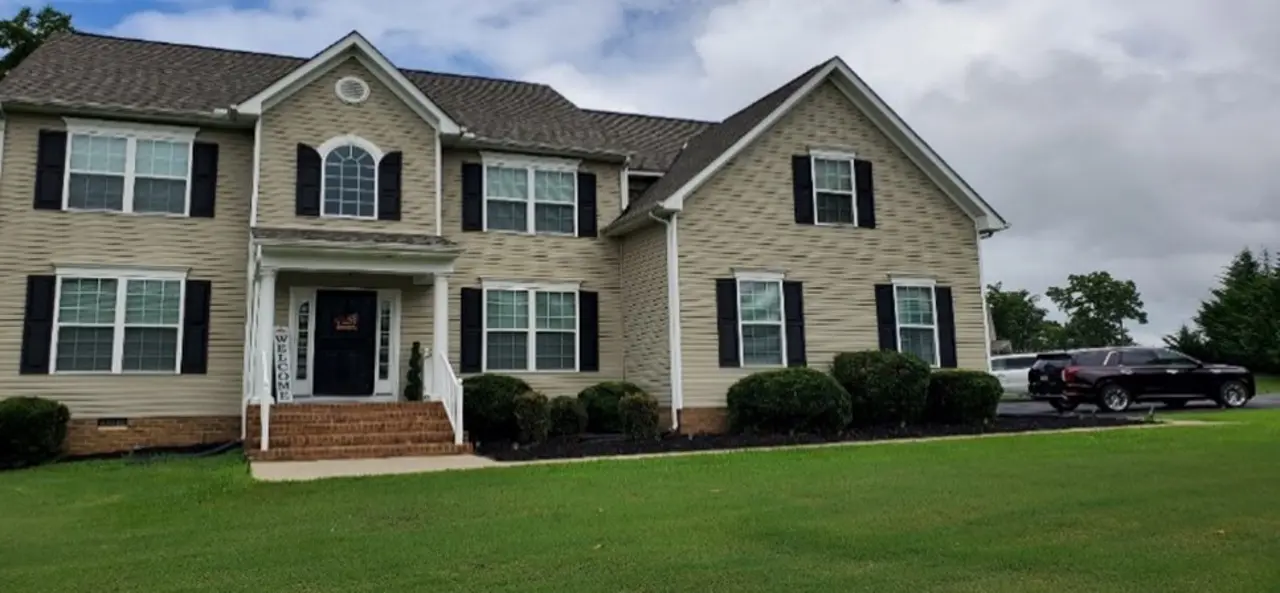 Image of a large, two-story house with beige siding, black shutters, and a well-maintained lawn. The home features a welcoming porch with stairs and a "Welcome" sign by the door. There is a driveway on the right side with a black SUV parked there. The sky appears partly cloudy, and the landscaping around the house is neatly trimmed.