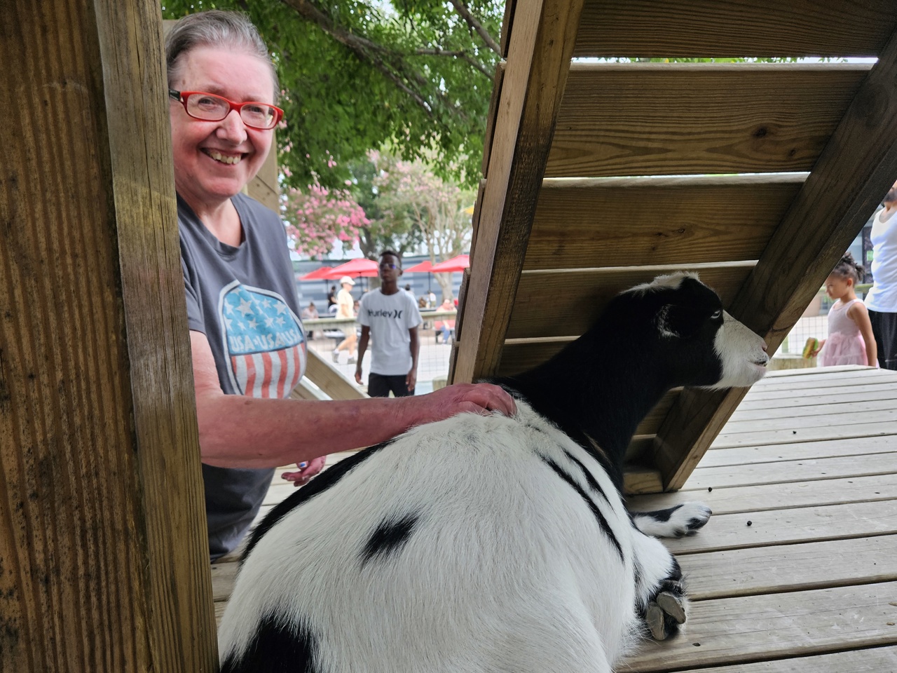 A woman grins widely while standing near a wooden structure and petting a black and white goat who is laying down. The scene is outdoors and other adults and children can be seen in the background.