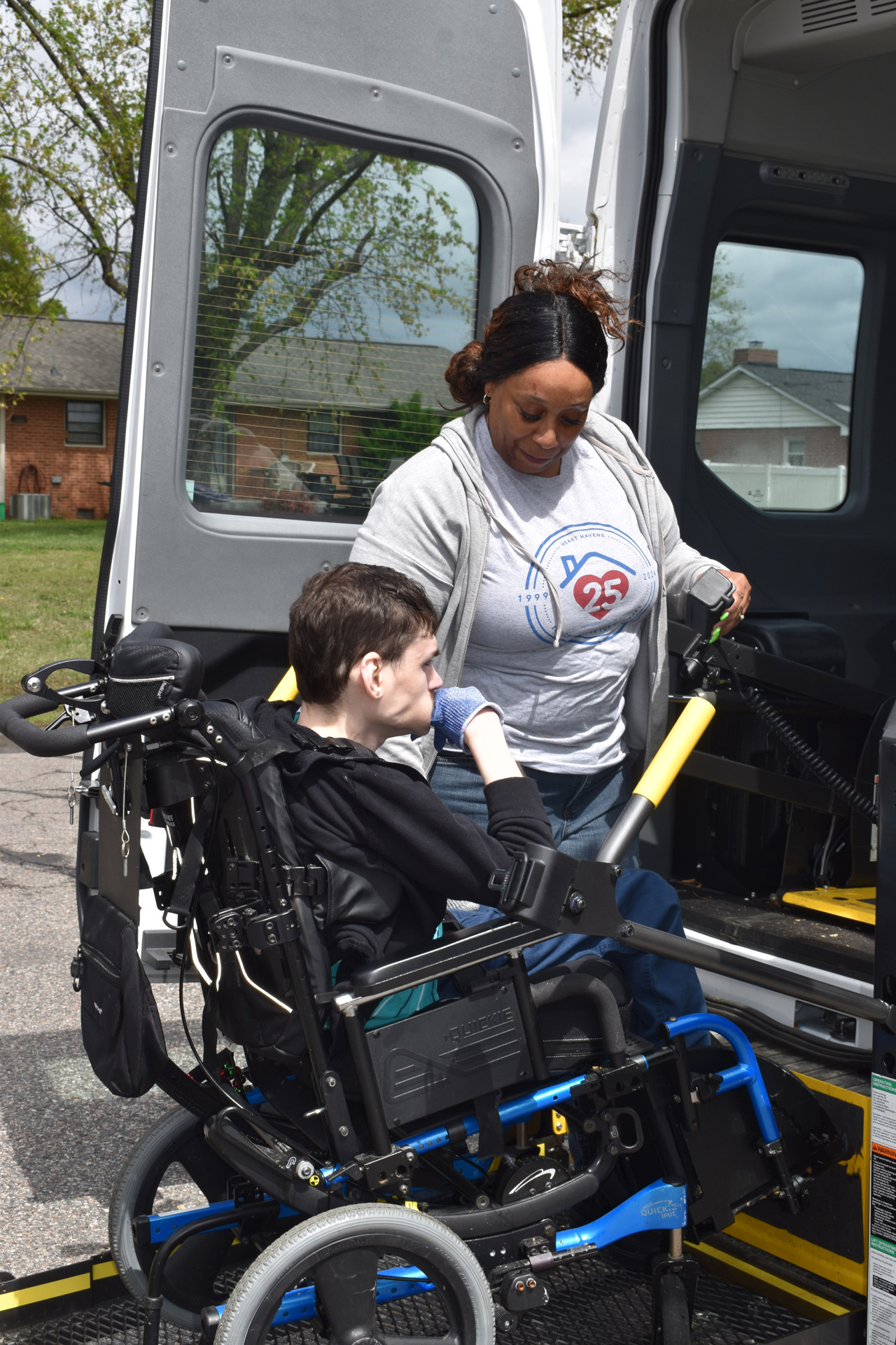 A Heart Havens staff member assists a resident in a wheelchair as they board a lift-equipped van. The staff member, wearing a Heart Havens 25th anniversary T-shirt and gray hoodie, stands beside the wheelchair lift, operating the controls. The resident is seated in a motorized wheelchair, wearing a black sweatshirt and blue gloves, and is being carefully supported onto the lift. The scene takes place outside on a partly cloudy day, with brick houses and green grass visible in the background.