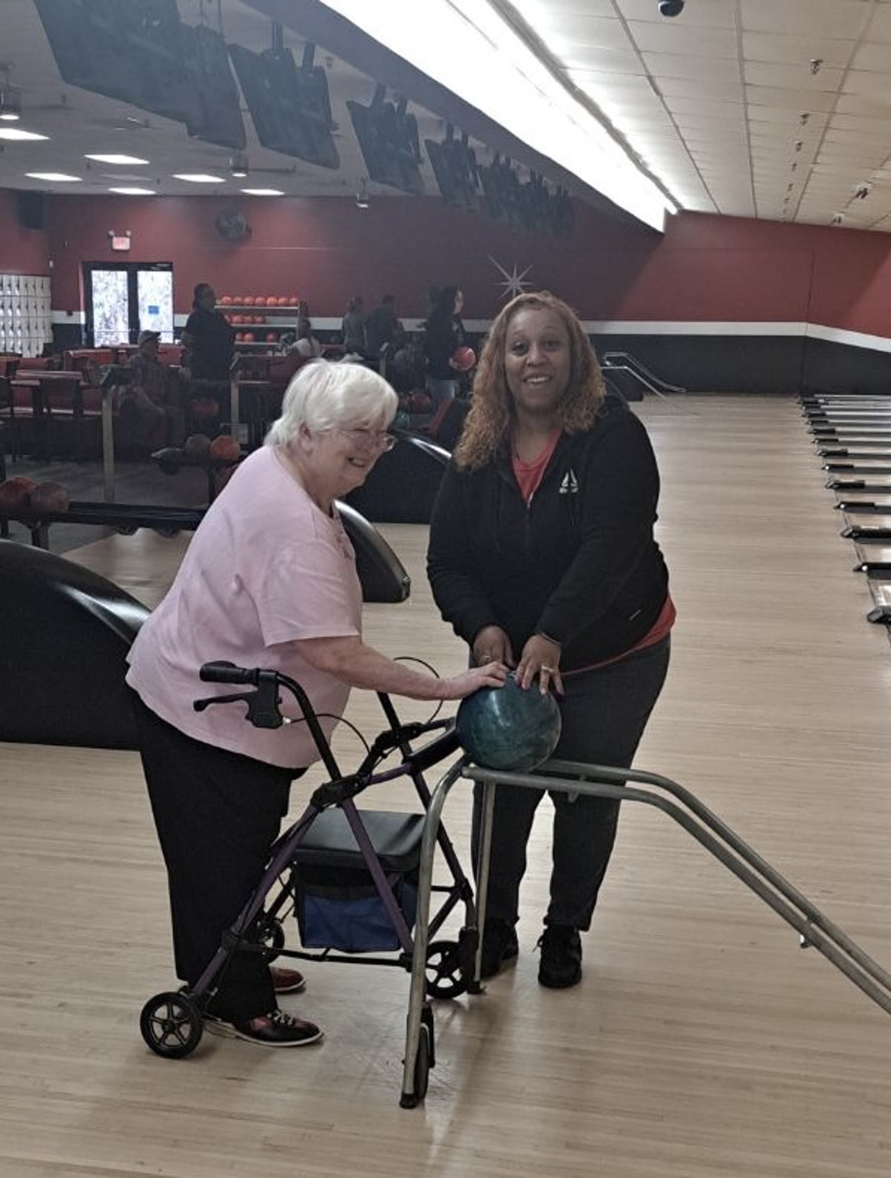 A woman with gray hair and another woman are enjoying a day at the bowling alley. The gray haired woman, wearing a pink shirt and using a walker, is smiling as she prepares to bowl with the assistance of a special ramp. The other woman, wearing a black hoodie, is helping her position the ball on the ramp. They are in a brightly lit bowling alley with several lanes visible in the background, along with other people bowling.