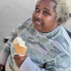 A woman with curly, short hair is sitting in a wheelchair outdoors, holding an ice cream cone and smiling. She is wearing a camouflage-patterned sweater. The background is blurred, focusing attention on her and her ice cream.
