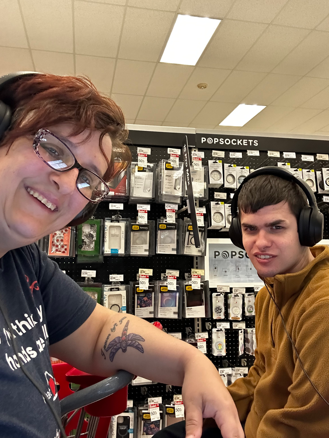 A smiling woman and a young man are posing for a selfie in what appears to be a retail store, specifically in the electronics section. The woman, wearing glasses and a black t-shirt with a tattoo on her left arm, holds the camera. The young man, dressed in a brown jacket, is smiling slightly and looking at the camera. Behind them, a display of various phone accessories, including PopSockets, is visible, indicating they are likely in a section dedicated to mobile phone gadgets and accessories.